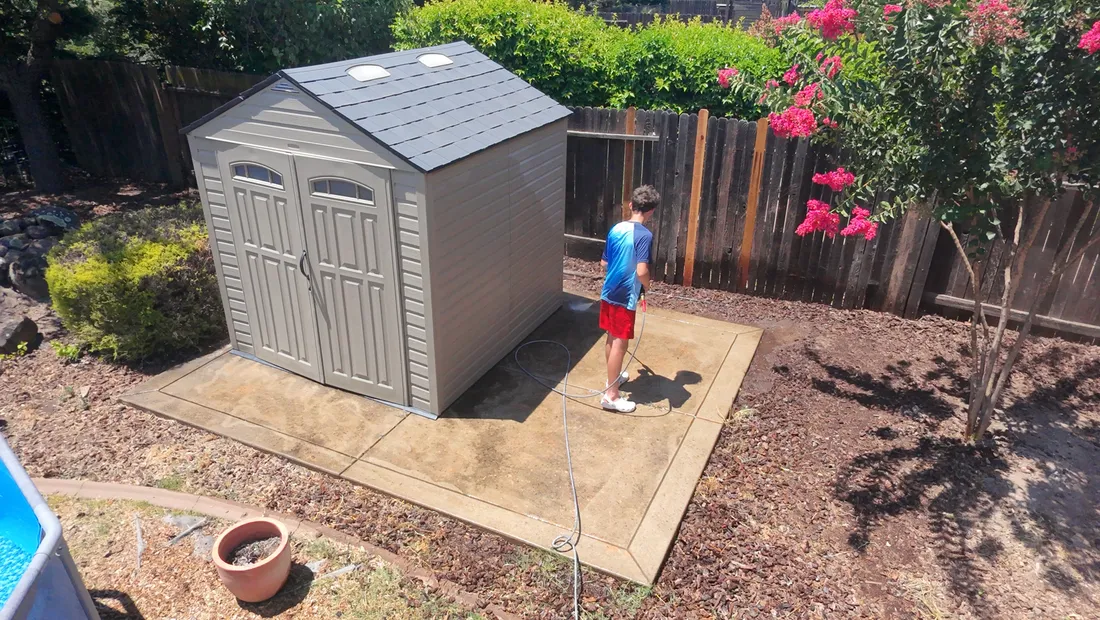 Concrete pad around a backyard shed being pressure washed, before cleaning