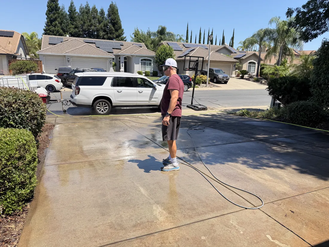 Crew member standing on a freshly washed driveway with pressure-washing equipment and hose staged near the vehicle, the wet concrete visibly darker than the dry pavement ahead