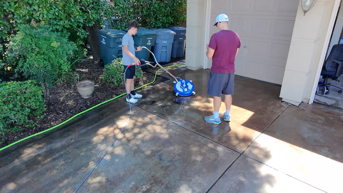 Two-person crew cleaning a concrete driveway apron near a garage door with a round surface cleaner attachment and hose