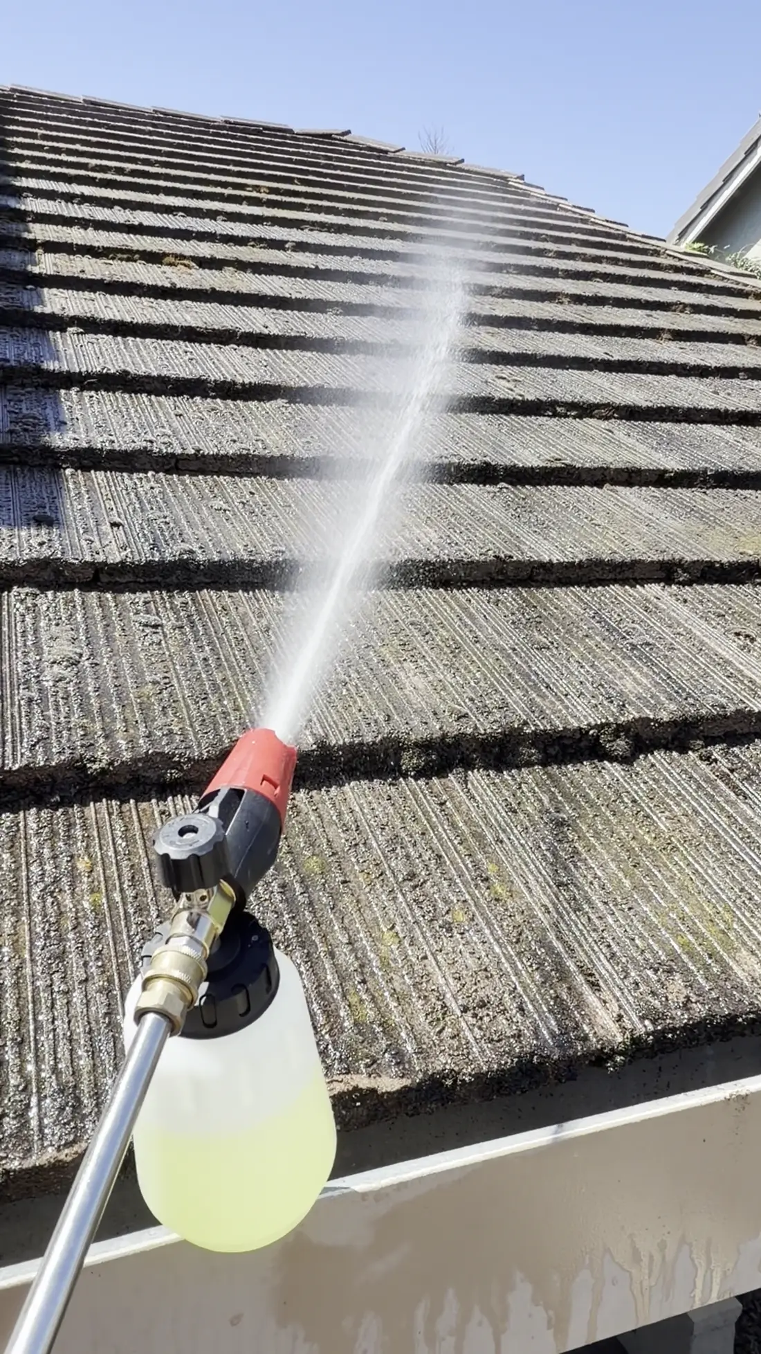 Cleaning solution misting from a low-pressure applicator onto flat concrete roof tiles darkened by lichen and moss