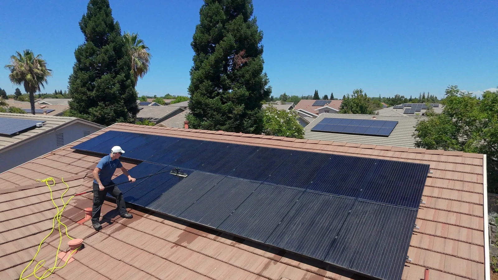 Cable Pressure Washing crew member standing on a tile roof cleaning a solar array with a water-fed pole brush, half the panels already washed