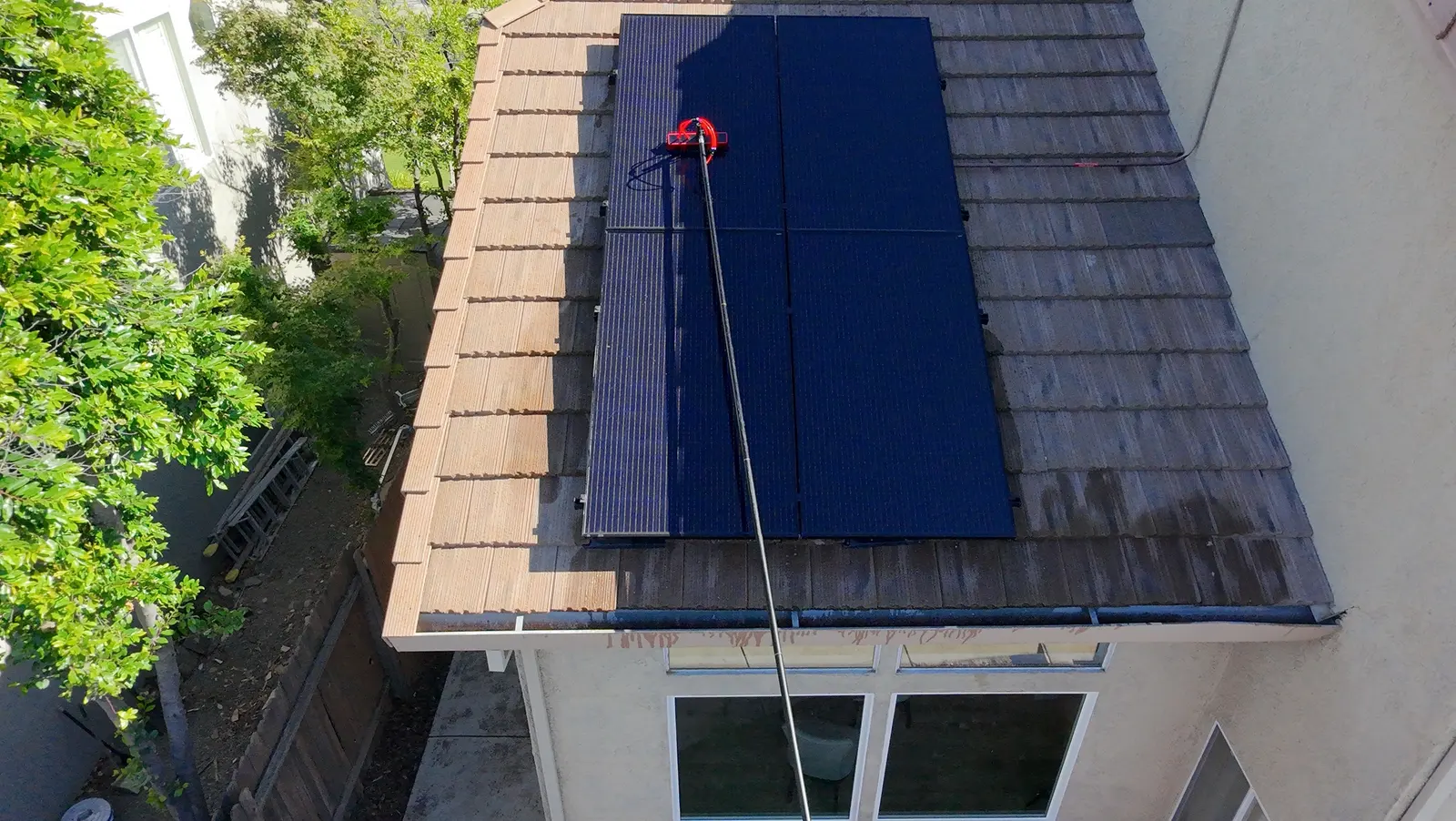 Water-fed pole with soft brush washing rooftop solar panels, the wet glass visibly clearing as grime lifts